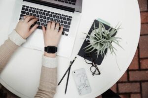 Is My Side Hustle A Hobby Or A Business? Woman Typing At Computer At Round Desk with Small Plant